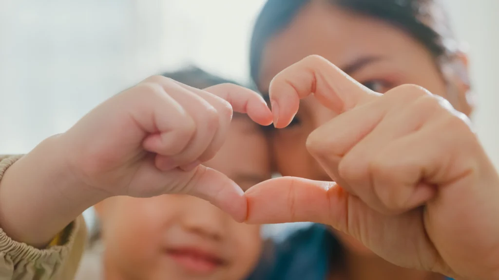 Closeup of young Asian family child making fingers heart shaped signs on sofa in living room at home.
